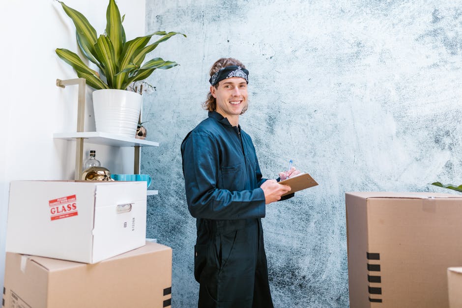 A young male cleaner with shoulder-length wavy hair, wearing a dark blue uniform and a patterned headband, is standing in a room with a textured light blue wall. He is smiling and holding a clipboard 